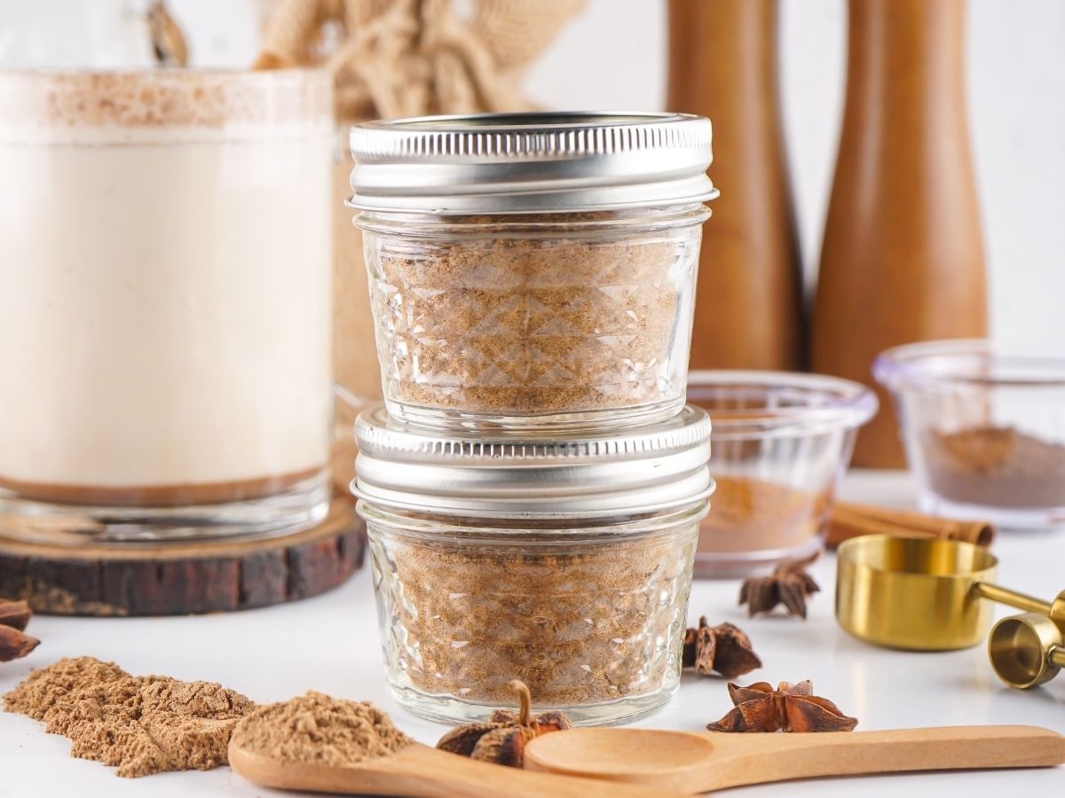 Two small glass jars with silver lids filled with a homemade aromatic seasoning blend stacked on a white surface.