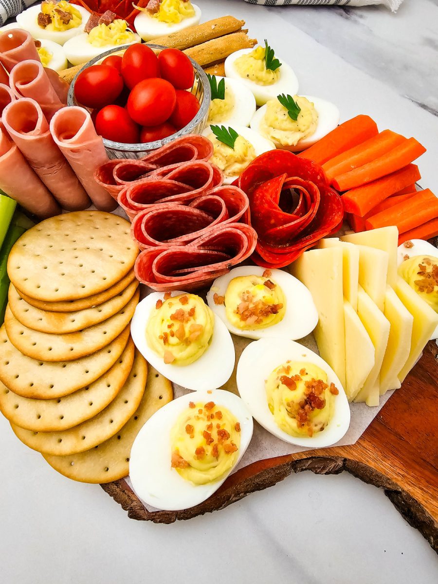 A close-up view of halved eggs topped with creamy yellow filling and crispy bacon bits, arranged on a wooden serving platter next to crackers and sliced cheese.