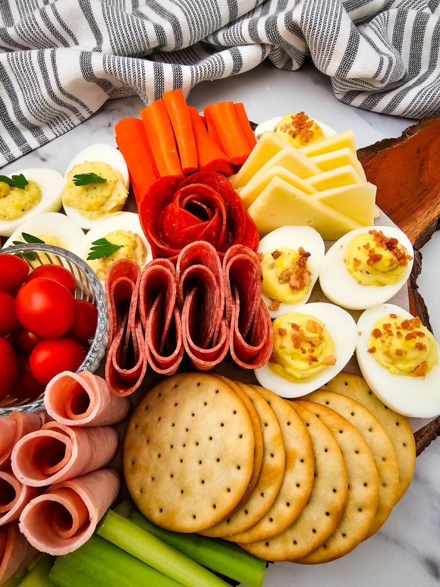 Close-up of a party tray featuring savory eggs with bacon crumbles, rolled ham slices, round crackers, and bright orange carrot sticks.