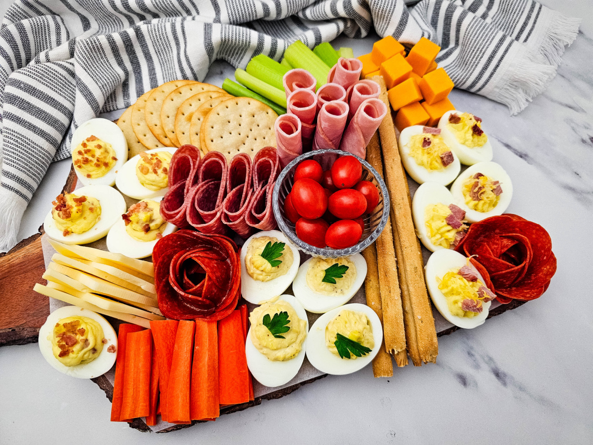A wide variety of appetizer snacks including eggs with herb garnishes, rolled deli meats, grape tomatoes in a glass bowl, and crunchy breadsticks.