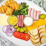 A large white oval platter filled with assorted deli meats, cheeses, and fresh toppings. This build-your-own party tray includes buttery croissants, crusty French bread slices, rolled ham, folded salami, and bowls of mustard and mayonnaise surrounded by pickles, onions, and tomatoes.
