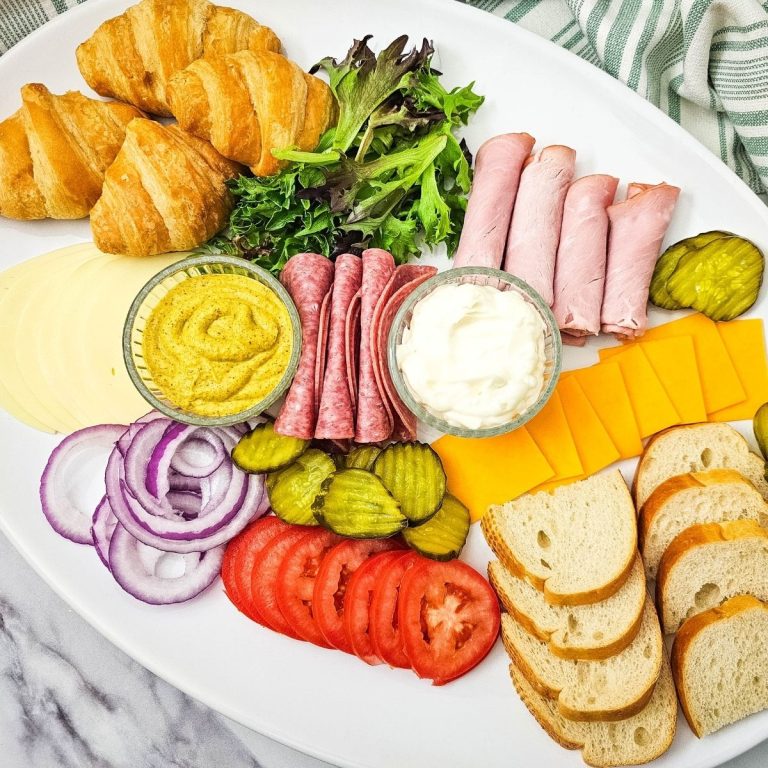 A large white oval platter filled with assorted deli meats, cheeses, and fresh toppings. This build-your-own party tray includes buttery croissants, crusty French bread slices, rolled ham, folded salami, and bowls of mustard and mayonnaise surrounded by pickles, onions, and tomatoes.