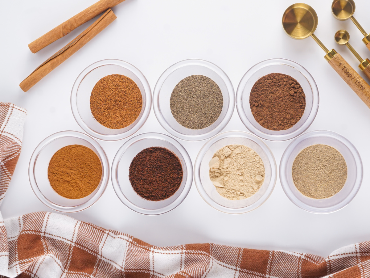 An overhead view of seven small glass bowls containing different ground spices like cinnamon and ginger arranged on a white surface with measuring spoons.