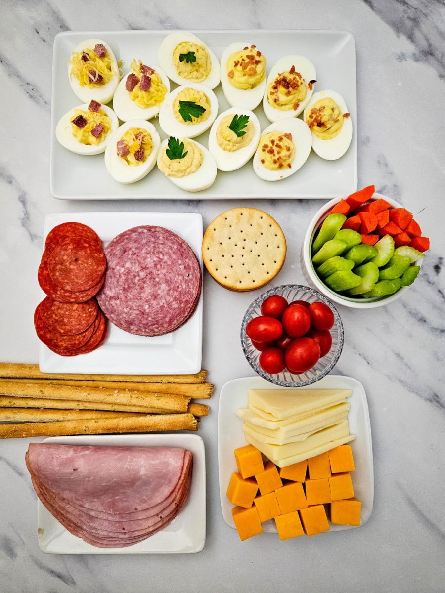 A flat lay of prepped ingredients on a marble surface featuring a tray of deviled eggs, sliced ham, salami, pepperoni, cheese, crackers, and vegetables.