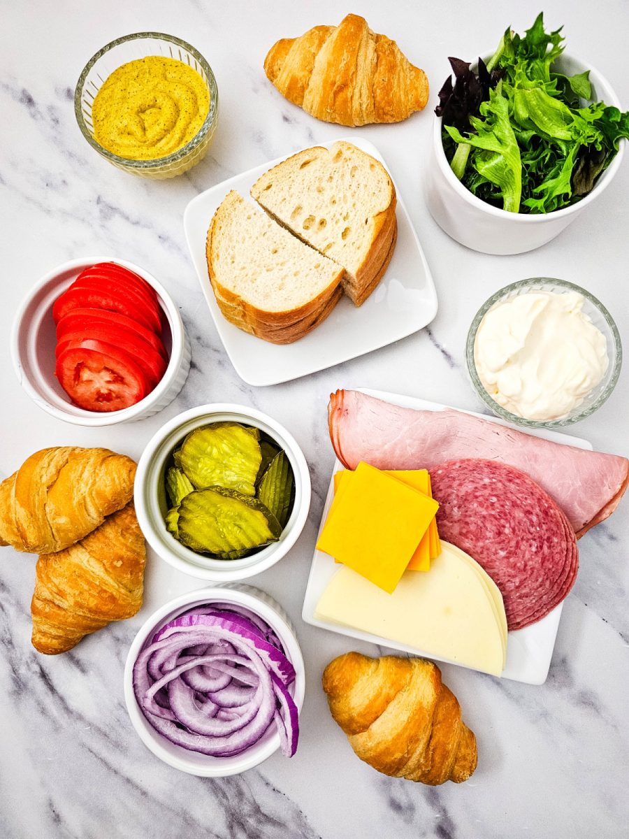 An overhead "flat lay" of all the components needed for a build-your-own sandwich station. Small white bowls hold pickles, red onions, tomatoes, and spring mix, surrounding plates of sliced deli meats, assorted cheeses, and golden croissants.