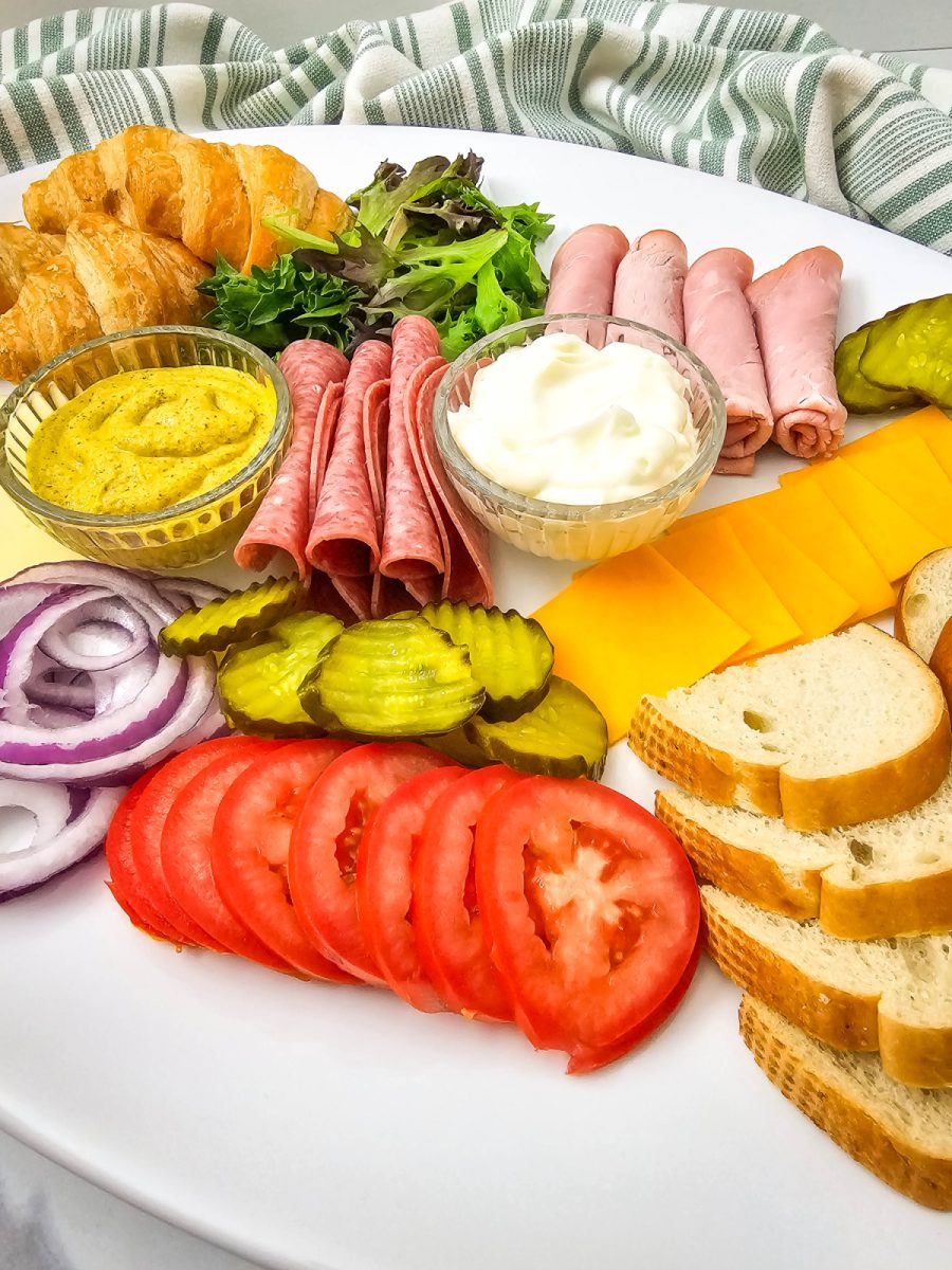 A close-up, angled shot of a gourmet sandwich spread. The image highlights the contrast between the flaky mini croissants and the vibrant red tomatoes and green lettuce, perfect for a light office lunch or casual home gathering.