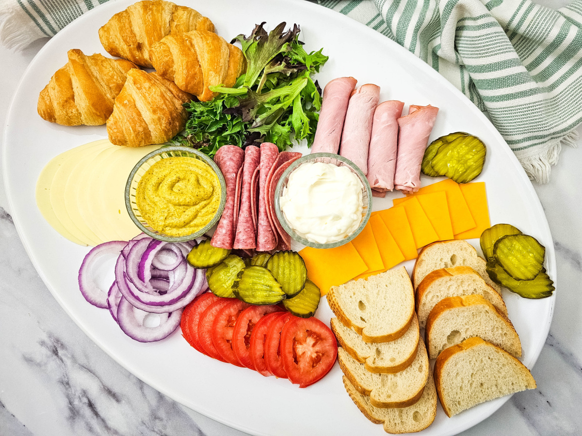 A high-angle view of a completed party platter featuring a variety of textures and colors. The arrangement includes stacks of crusty bread, rolled deli meats, sliced cheeses, and fresh garnishes like pickles and onions organized for easy grazing.