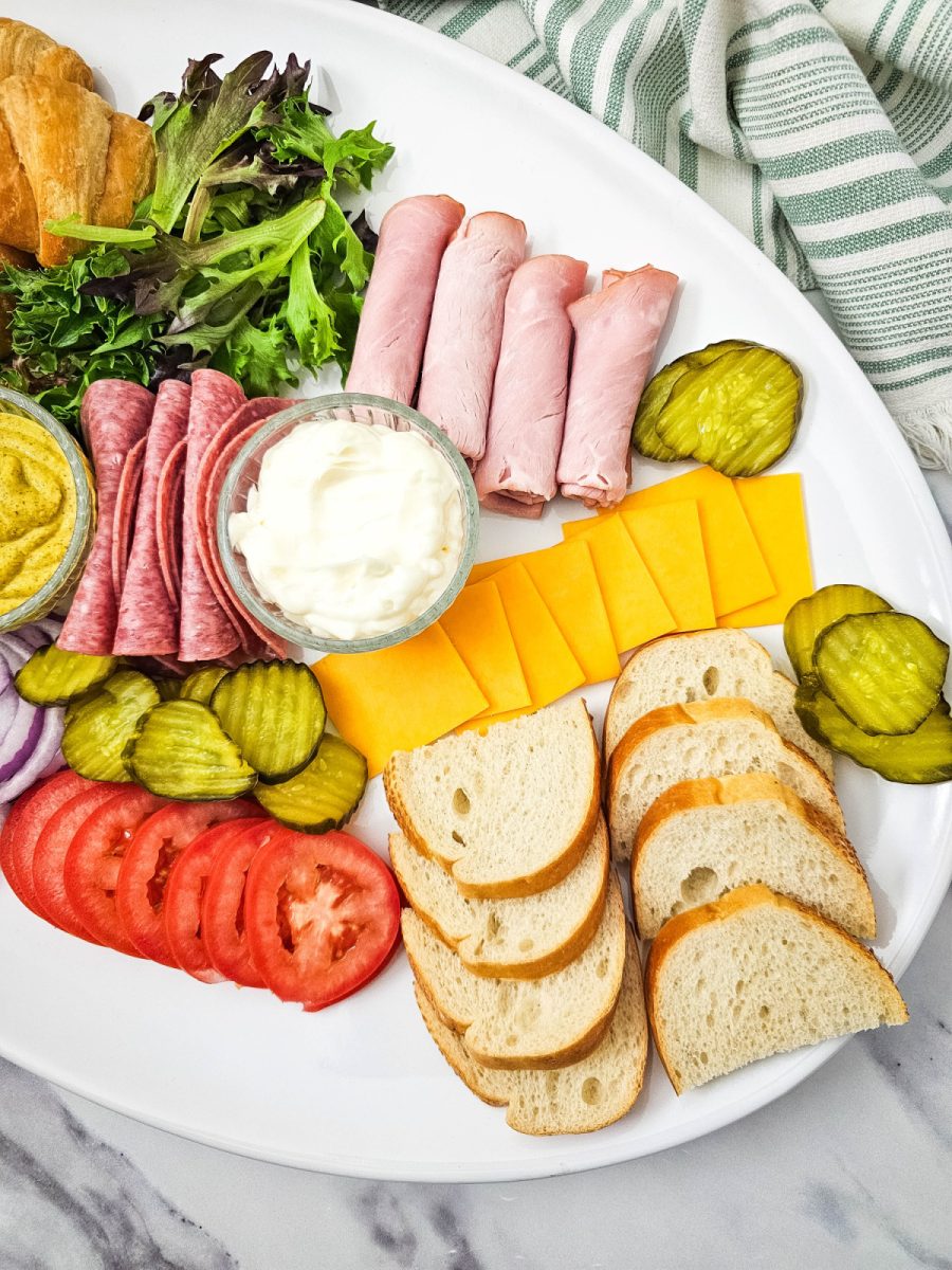 A top-down perspective of a meat and cheese tray focused on the right side of the arrangement. It shows the orderly rows of cheddar cheese slices, crusty bread rounds, and ham rolls next to a striped kitchen towel.