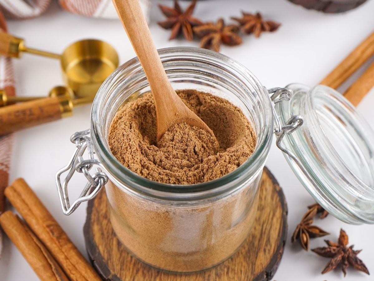 A wooden spoon dipped into a clear glass jar of a homemade spice powder surrounded by whole cinnamon sticks and star anise.