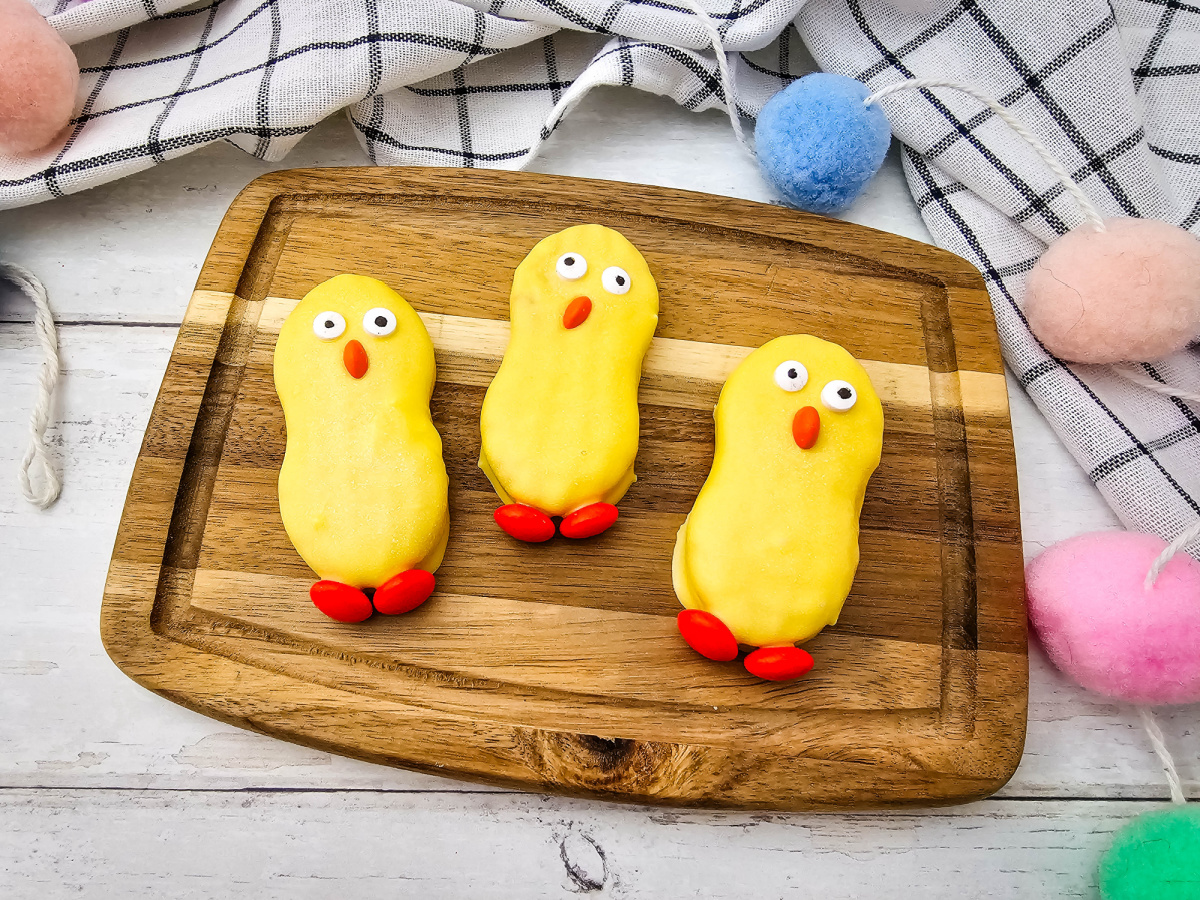 hree yellow-dipped peanut butter cookies decorated with candy eyes, an orange beak, and orange candy feet, arranged on a small wooden cutting board.