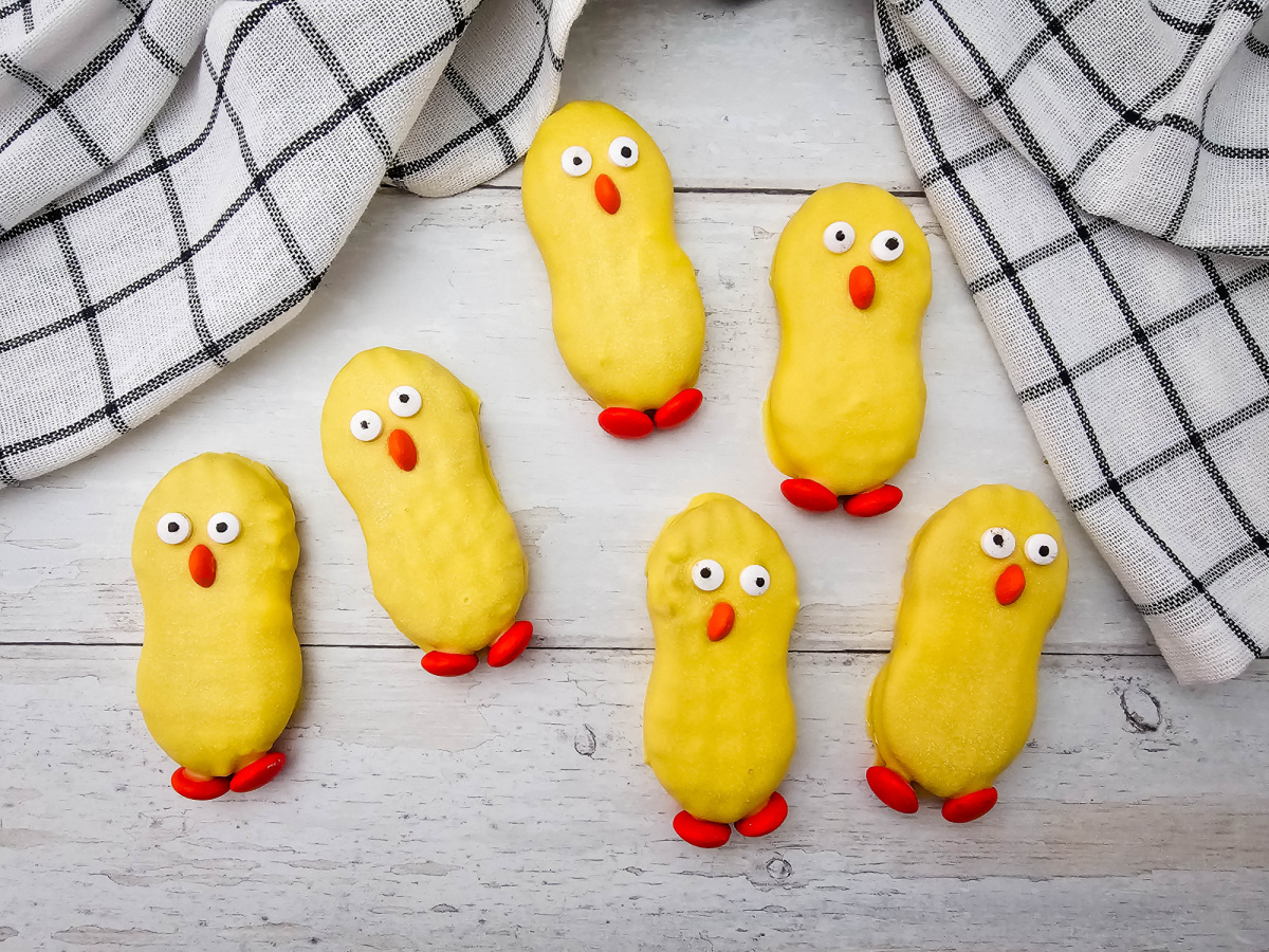 Six handmade yellow chick cookies scattered on a white wooden surface next to a black and white checkered kitchen towel.