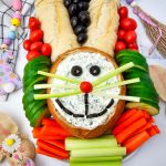 A bright holiday appetizer tray with a round dip bowl face, bread ears, and organized rows of crisp carrots and garden-fresh vegetables