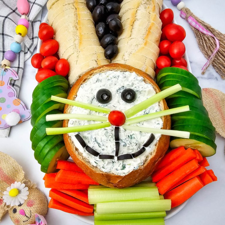 A bright holiday appetizer tray with a round dip bowl face, bread ears, and organized rows of crisp carrots and garden-fresh vegetables
