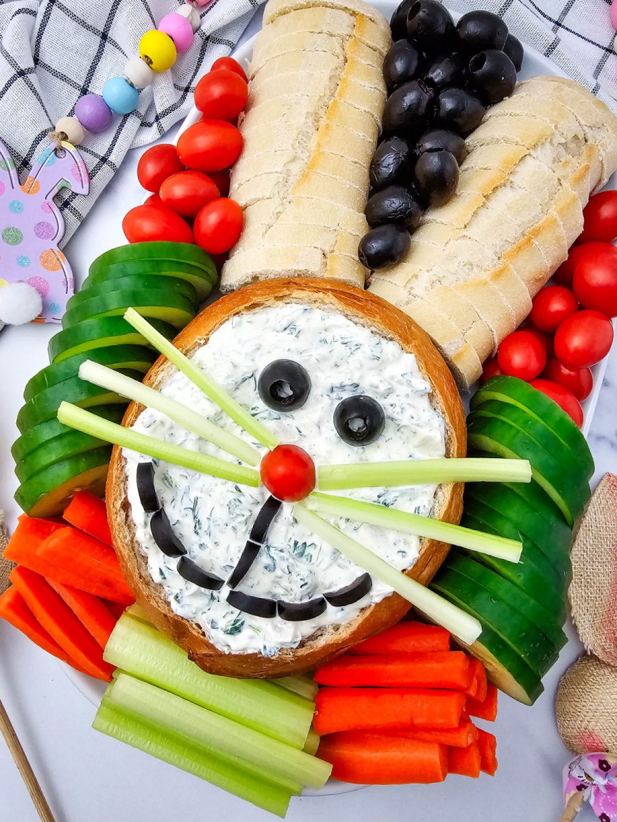Close up of a sourdough loaf hollowed into a bowl and filled with herb dip, decorated with garden vegetables to create a whimsical animal face.