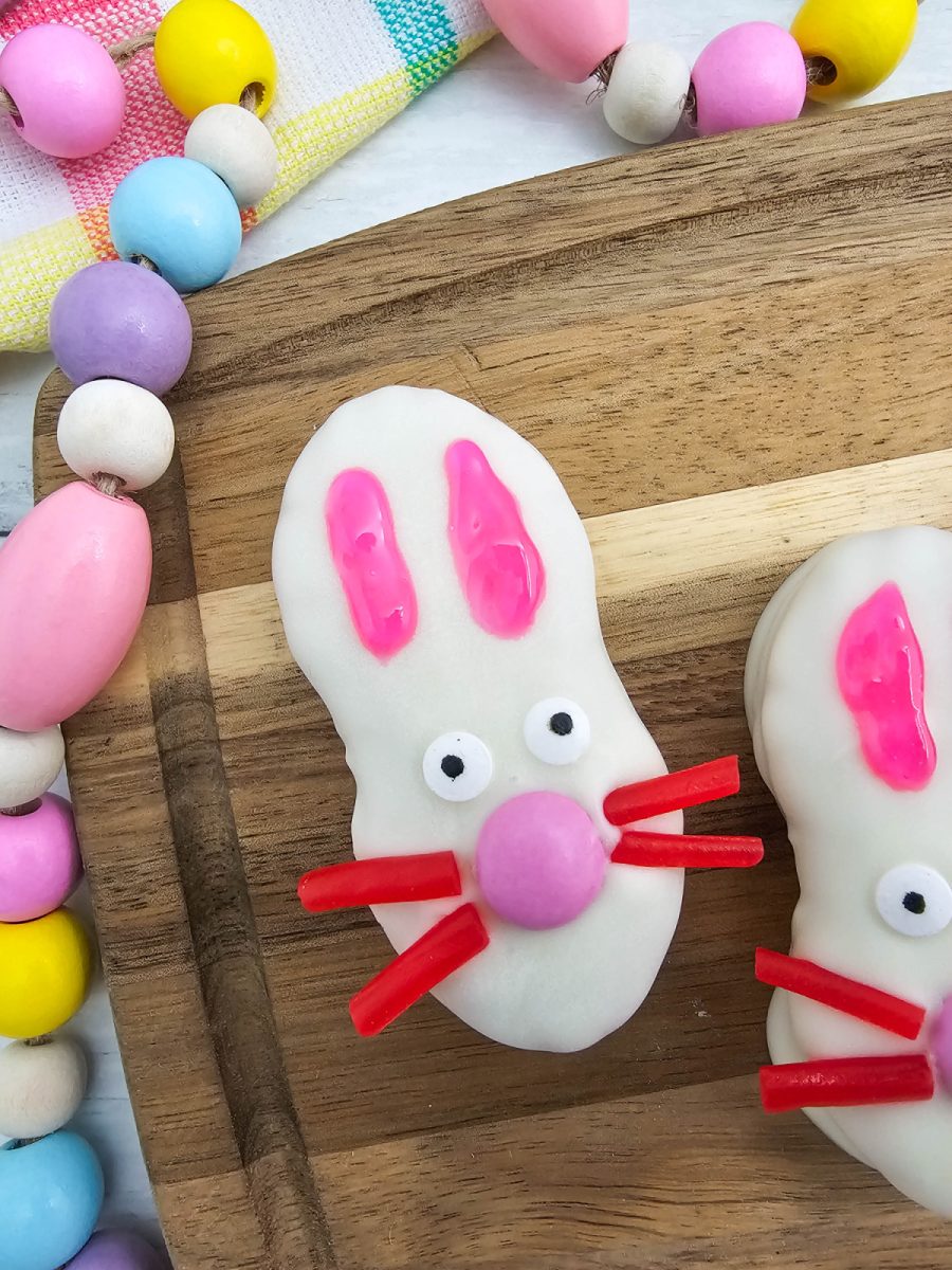 A detailed close-up of a single Nutter Butter bunny cookie showing the texture of the white chocolate coating, red licorice whiskers, and small candy eyes.