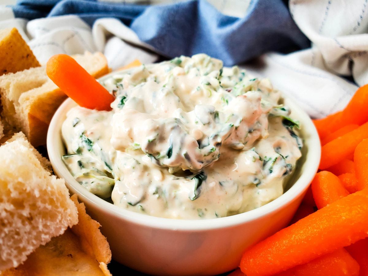 Close up of a creamy dip recipe in a white bowl served with a baby carrot and crusty bread.