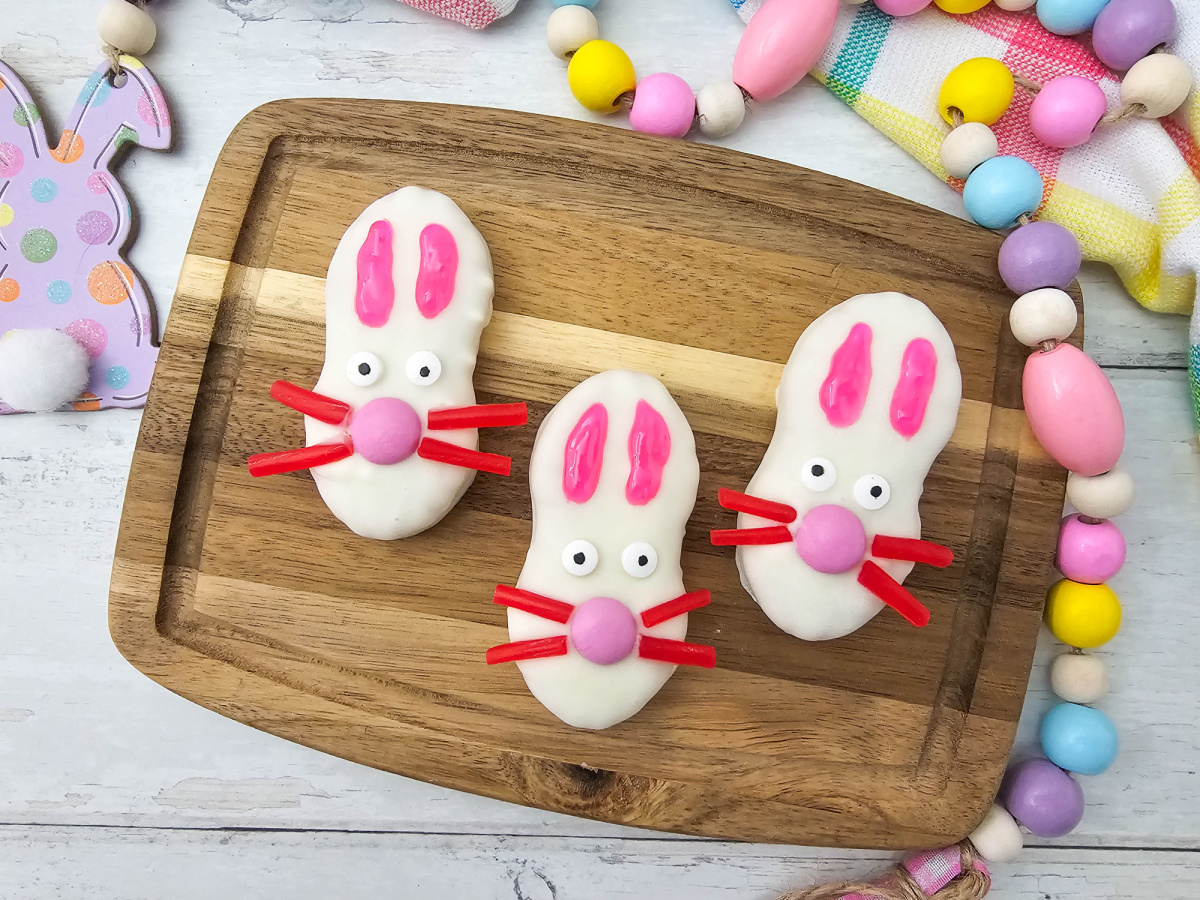 A high-angle shot of three Nutter Butter bunny cookies on a wooden board surrounded by a colorful pastel wooden bead garland and an Easter bunny decoration.
