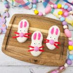 The primary recipe photo showing three adorable white-coated bunny cookies arranged on a wooden board with festive Easter decor and a plaid napkin in the background.