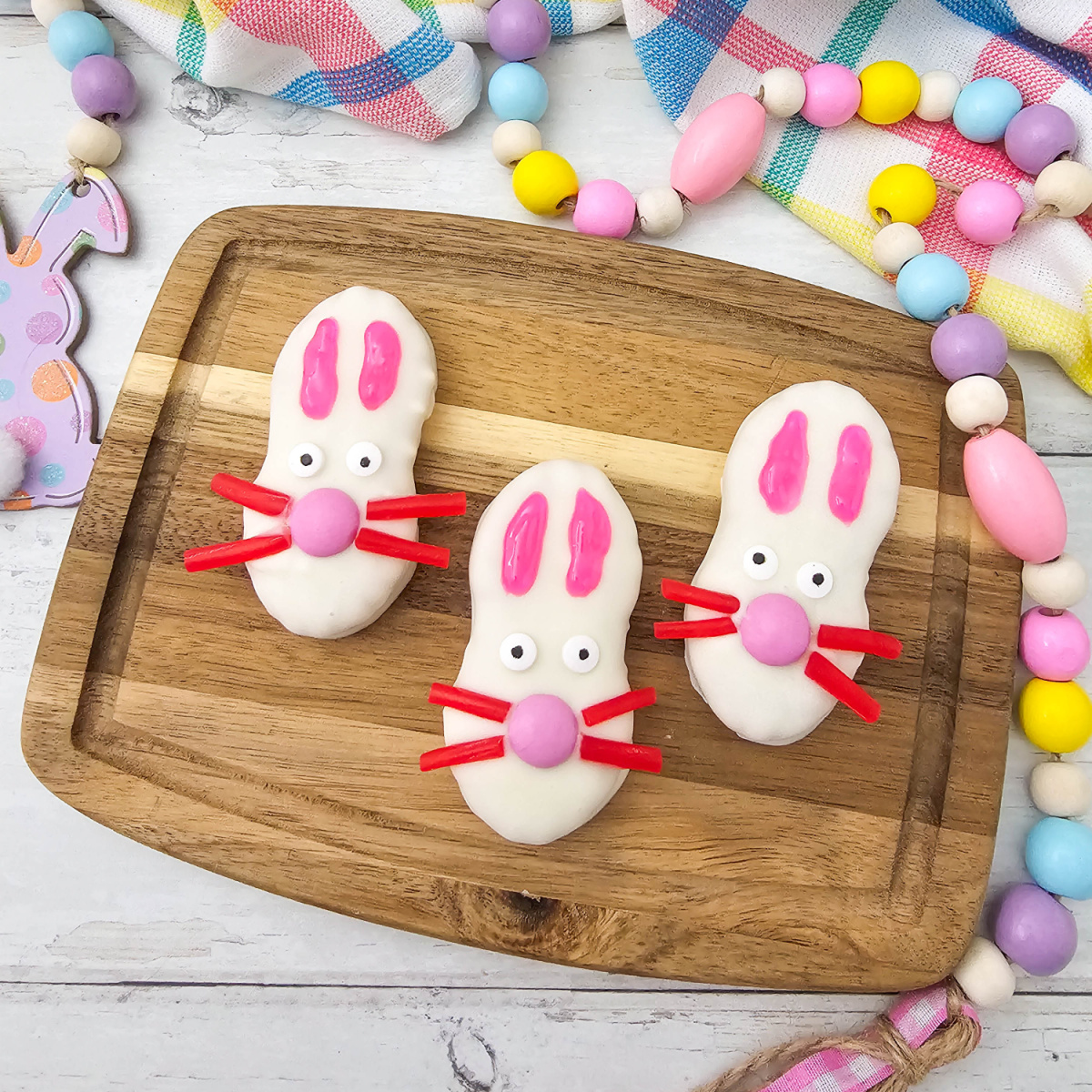 The primary recipe photo showing three adorable white-coated bunny cookies arranged on a wooden board with festive Easter decor and a plaid napkin in the background.