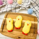 A top-down view of three adorable yellow chick cookies on a wooden board, framed by colorful pastel pom-pom garland and a tea towel.