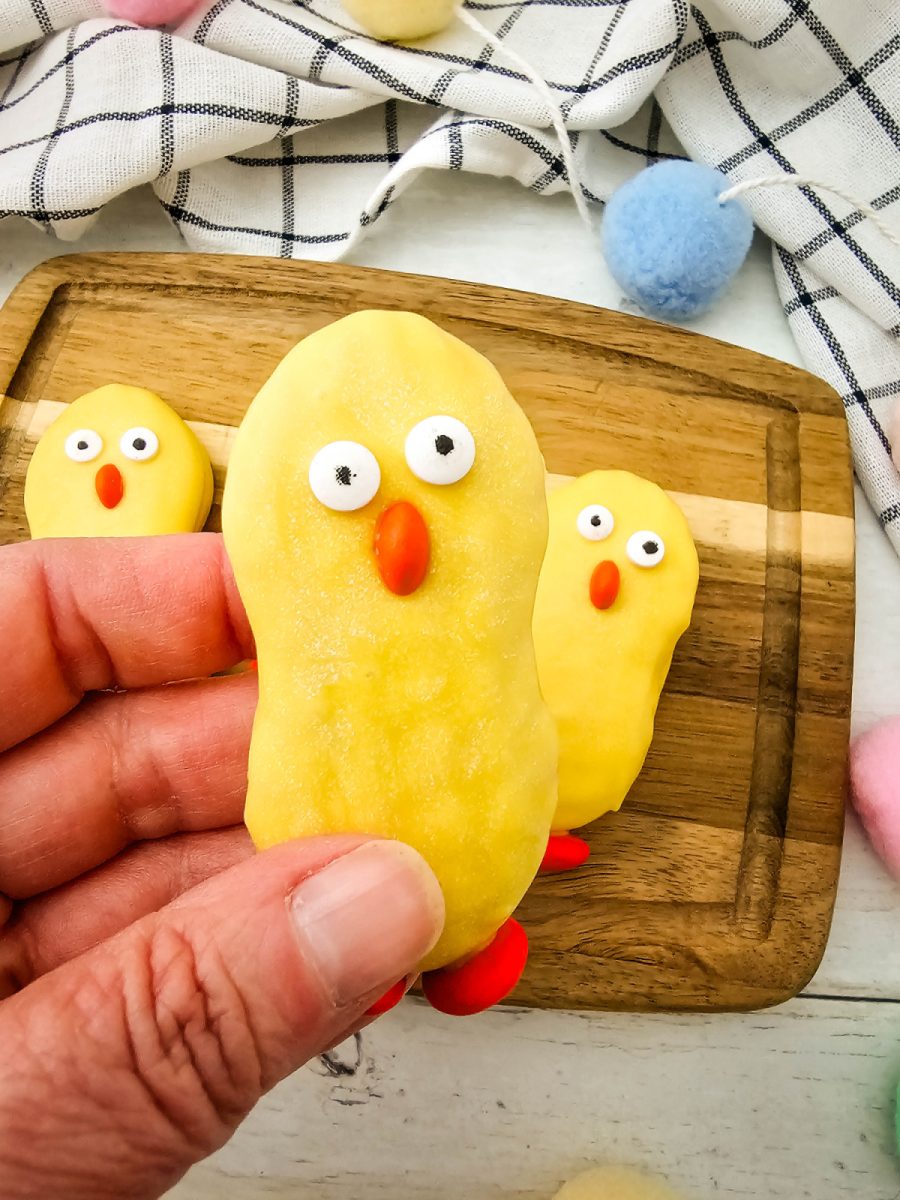 A person’s hand holding up one finished yellow chick cookie to show the candy eye and beak details, with more cookies on a board in the background.