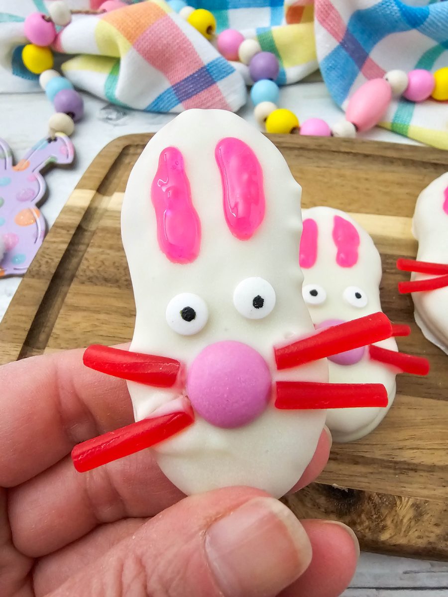 A person’s hand holding up a finished Nutter Butter bunny cookie to show its size and the playful details of the candy face and pink icing ears.