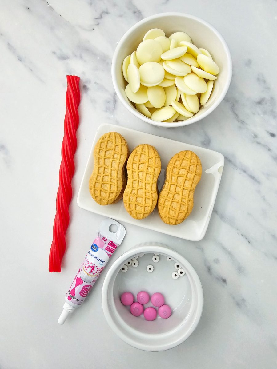 A flat-lay image of the ingredients needed for the recipe including Nutter Butter cookies, white chocolate melting wafers, a tube of pink decorating gel, pink M&Ms, candy eyes, and a red licorice twist.