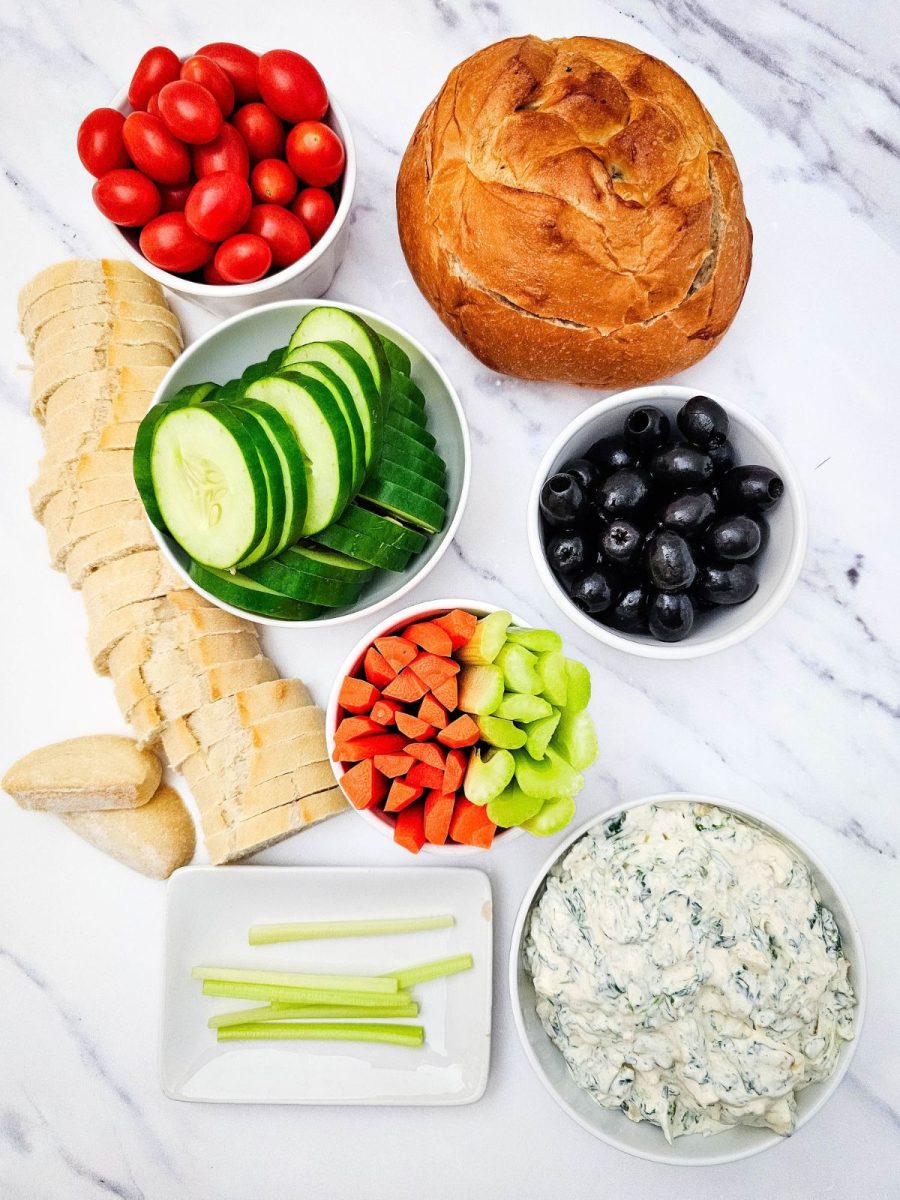 A collection of fresh components for a festive platter including a bread loaf, baguette, cherry tomatoes, cucumbers, olives, carrots, celery, and spinach dip.
