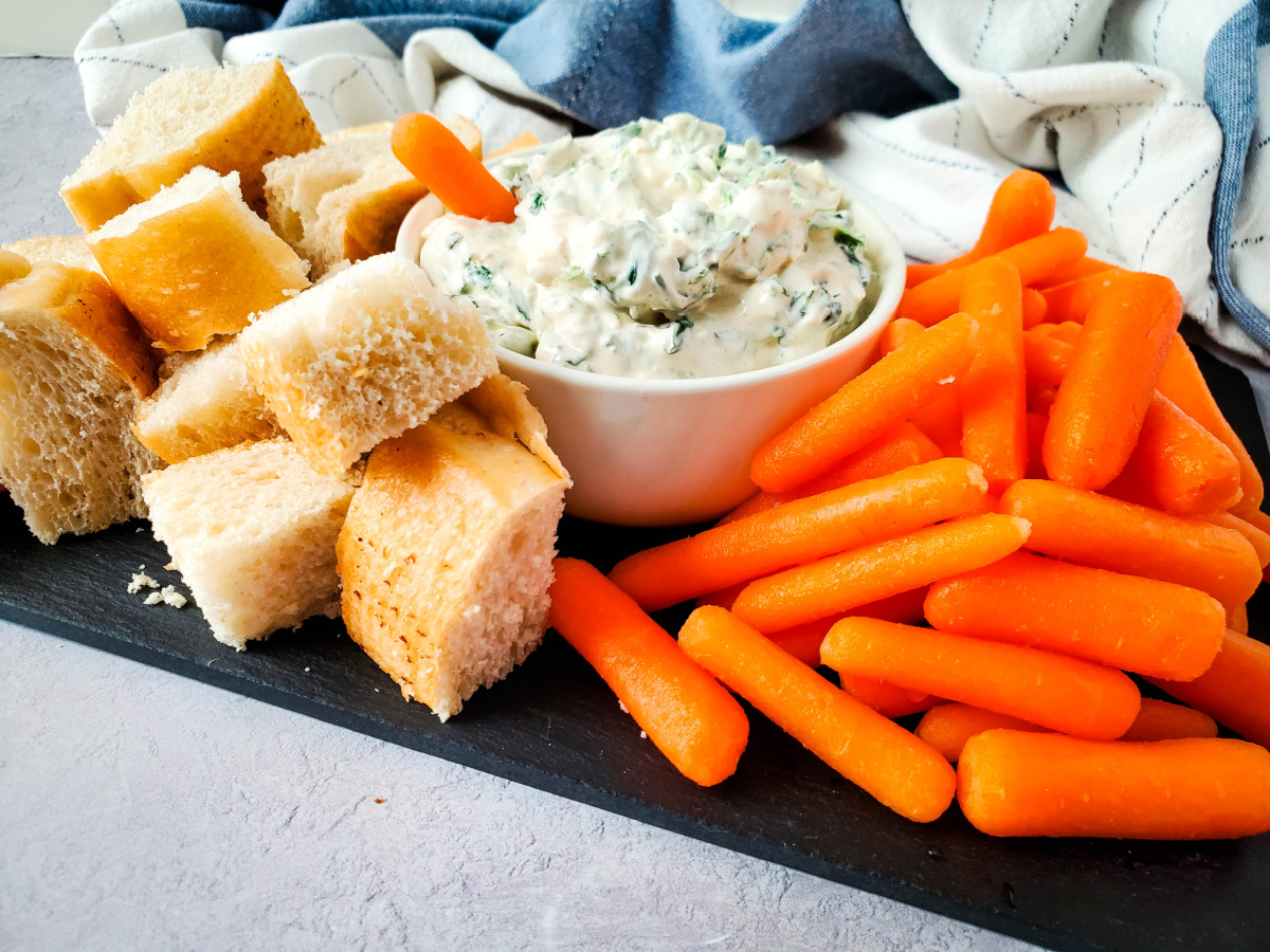 A large serving board with a white bowl of homemade spinach dip served alongside crisp baby carrots and bread cubes.