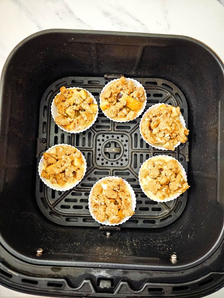 Overhead view of six unbaked French toast muffins with crumb topping in silicone liners arranged in a black air fryer basket.