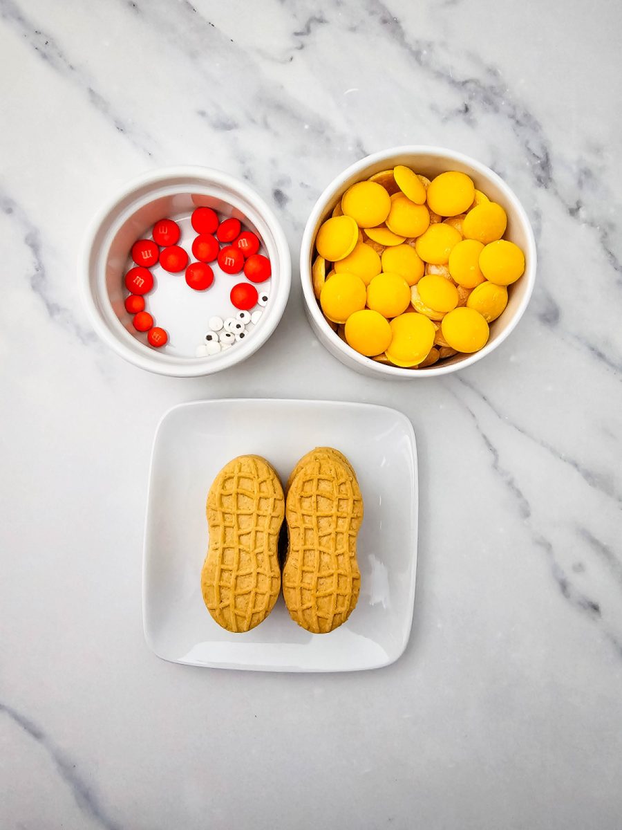 Overhead shot of the ingredients needed for the project, including a white plate with two peanut-shaped sandwich cookies and two small bowls containing yellow candy wafers, orange chocolate candies, and tiny candy eyes.