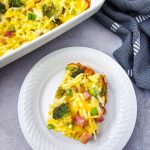 A vibrant close-up of a Ham and Broccoli Breakfast Casserole serving on a white plate with the full baking dish and a blue kitchen towel in the background.