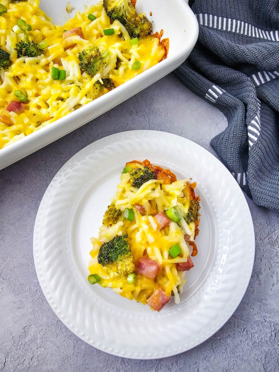 Top-down view of a savory egg bake with ham and broccoli, showing a fresh slice served on a white scalloped plate next to the baking dish.