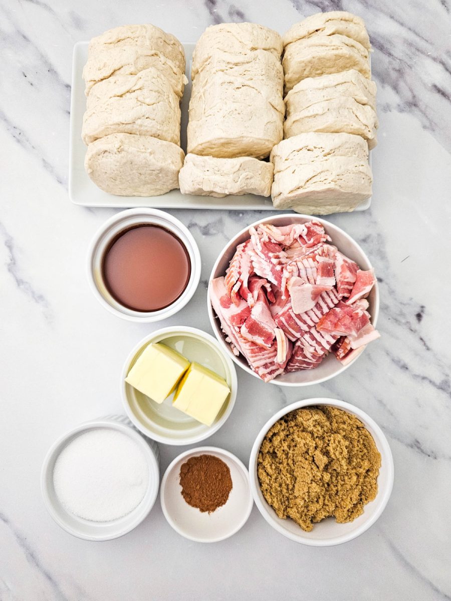 A flat lay of all necessary ingredients including canned biscuits, raw bacon, maple syrup, butter cubes, white sugar, cinnamon, and brown sugar.