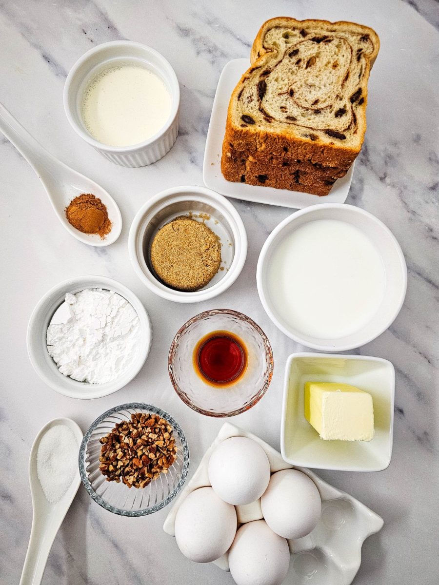 An overhead view of ingredients for French toast muffins including cinnamon raisin bread, eggs, milk, cream, butter, brown sugar, pecans, flour, and vanilla.