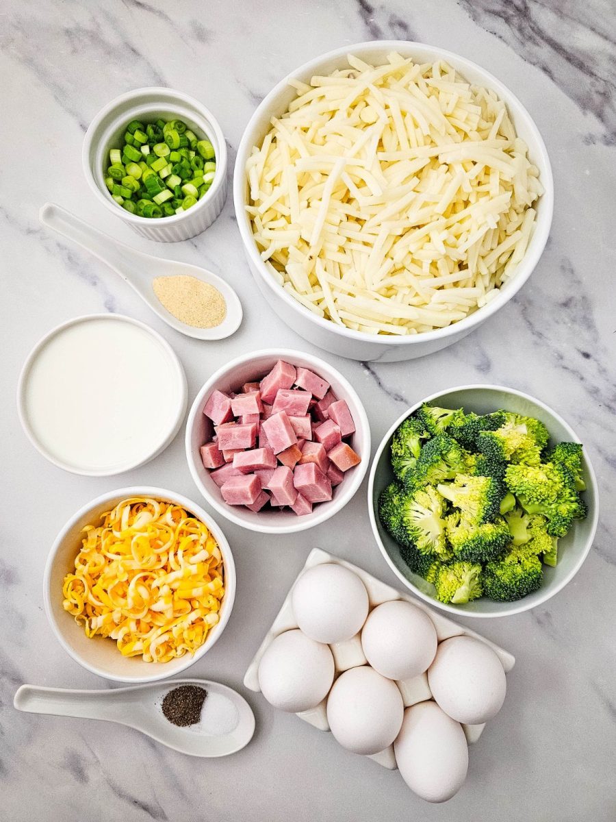 Top-down view of recipe ingredients including shredded potatoes, diced ham, fresh broccoli florets, eggs, milk, cheese, and seasonings in white bowls.