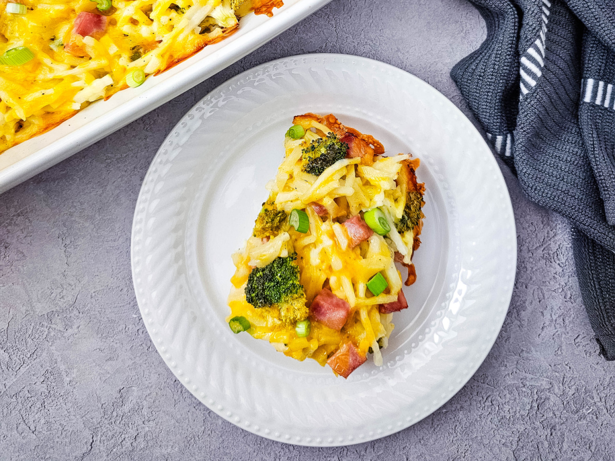 A single serving of a potato and egg bake with ham and broccoli, highlighting the melted cheese and crispy edges, served on a textured white plate.