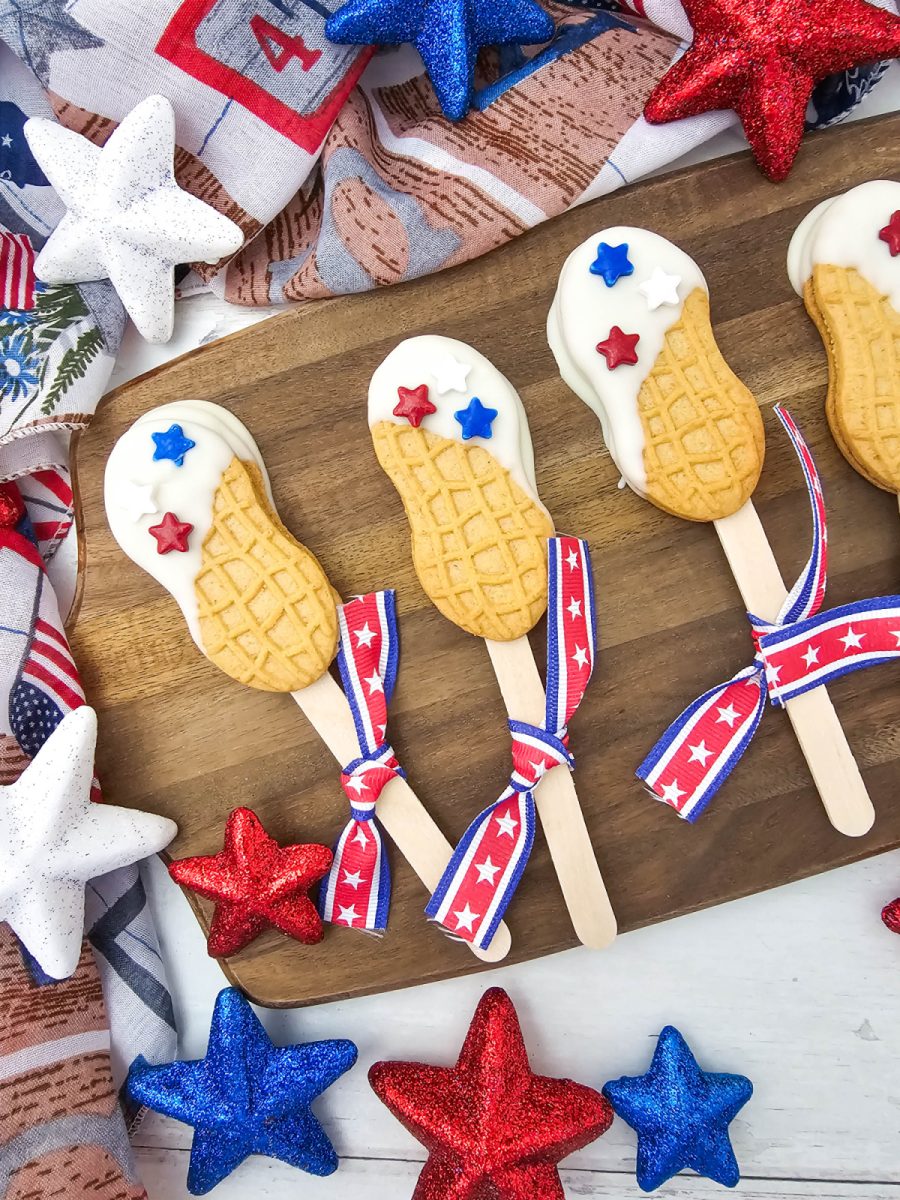 A close-up shot of several patriotic nutter butter cookie pops on wooden sticks decorated with white candy coating, bright red stars, and royal blue star sprinkles. The festive holiday treats sit on a rustic wooden tray with a patriotic patterned cloth background.