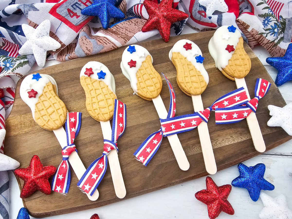 Five decorated red, white, and blue nutter butter cookie pops arranged in a row on a wooden serving board. Each cookie pop is partially dipped in white chocolate frosting, adorned with patriotic star sprinkles, and finished with a red, white, and blue star ribbon tied to the stick.