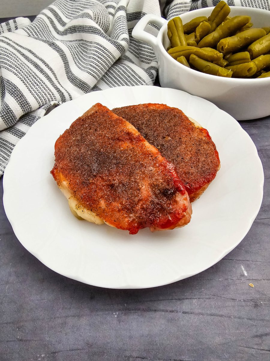 Two fully cooked, seasoned pork chops served on a white plate with a side dish of green beans and a striped kitchen towel.