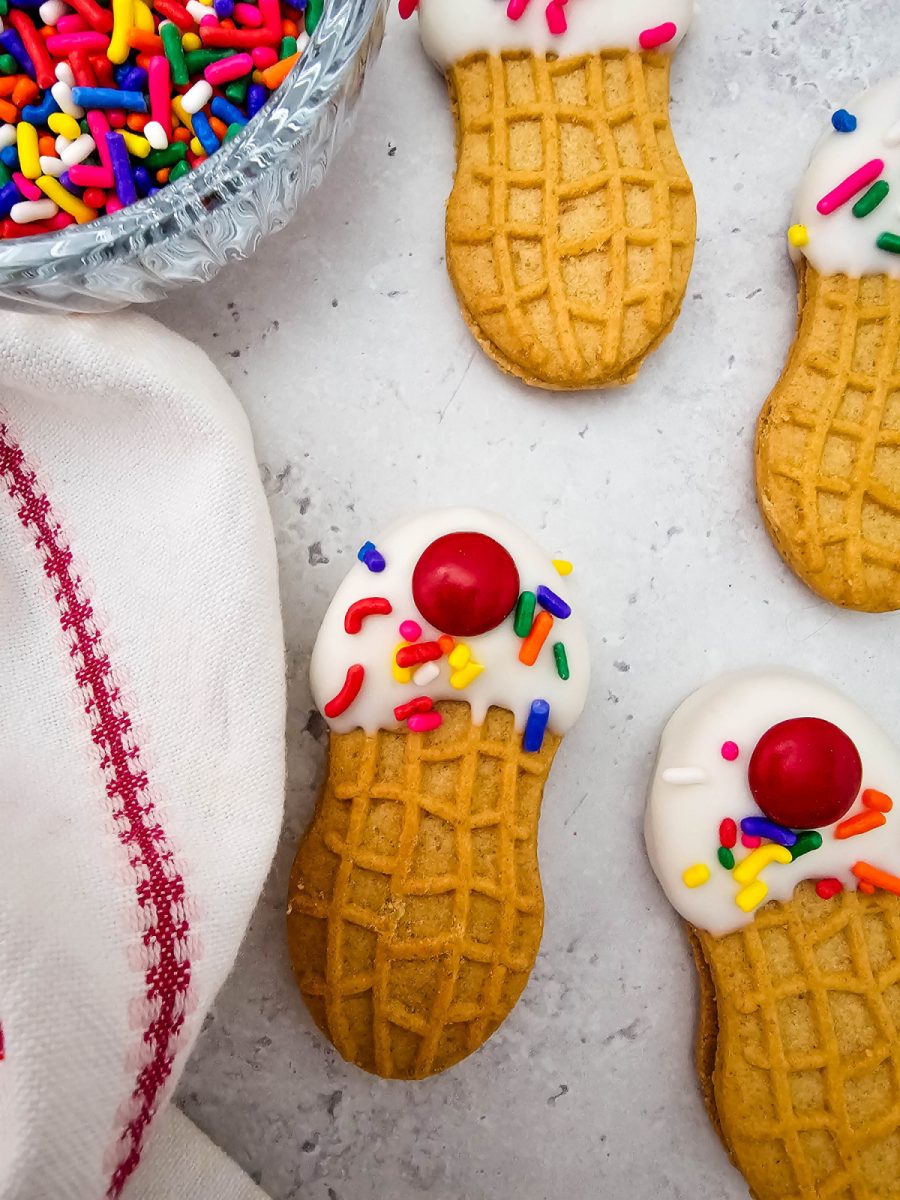 A vertical close-up view focusing on two peanut butter cookies decorated with a white candy coating shell, rainbow sprinkles, and a red candy top, positioned next to a bowl of sprinkles and a white cloth with red stitching.