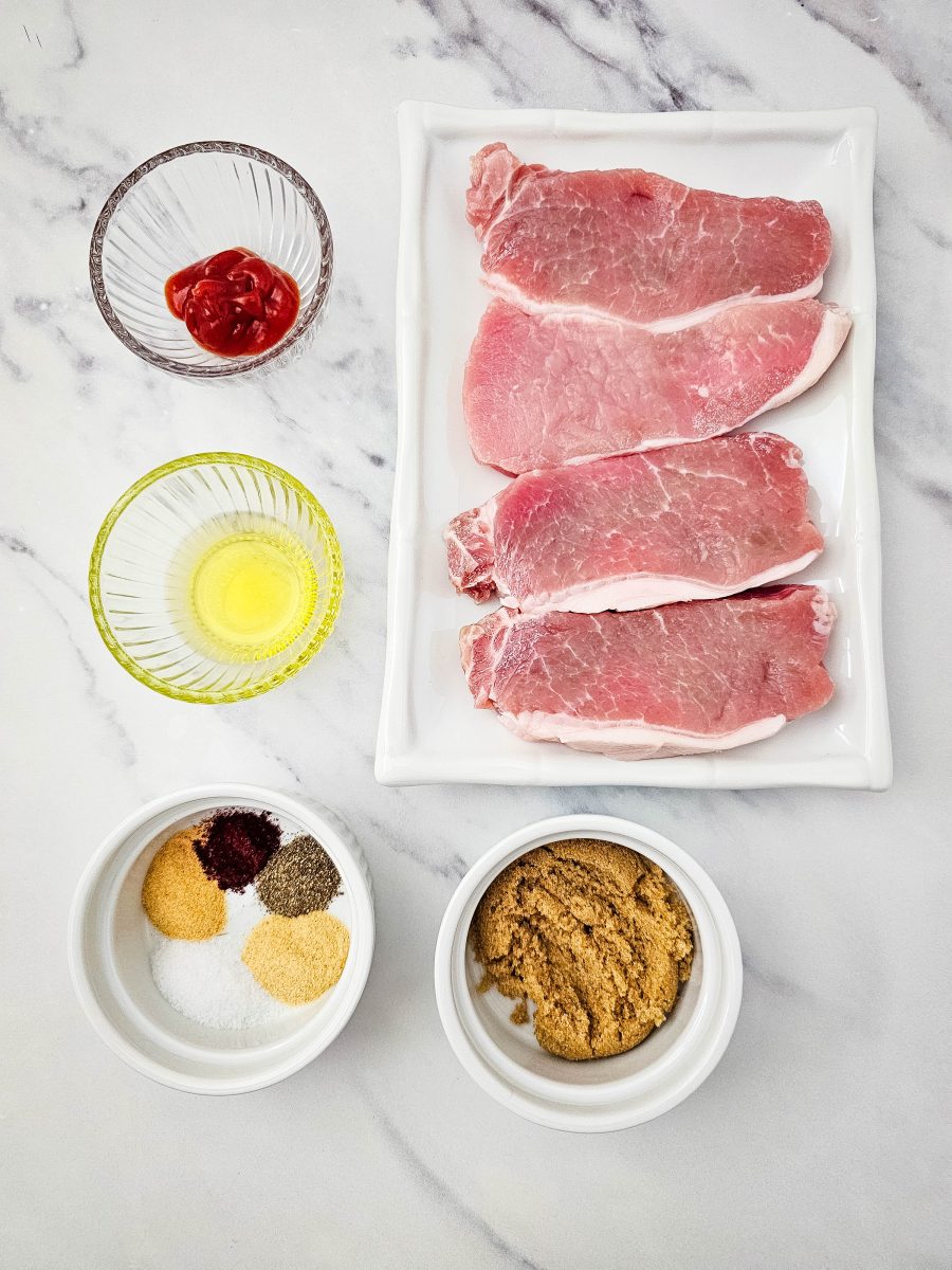An overhead view of raw pork chops on a rectangular platter surrounded by small bowls of olive oil, ketchup, brown sugar, and various dry seasonings.