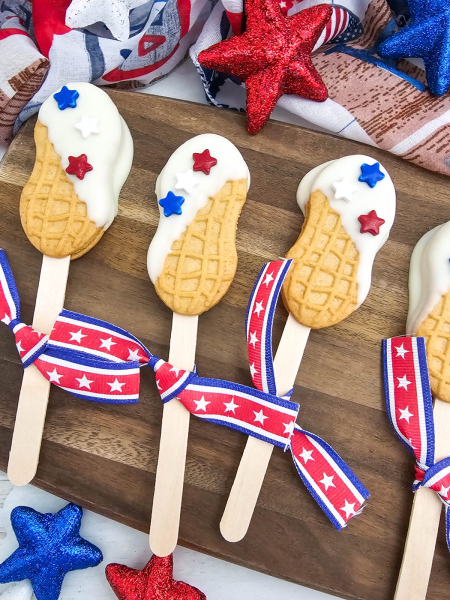 A beautifully styled flat lay of red, white, and blue nutter butter cookie pops mixed with festive 4th of July party decor. The dipped cookies feature white frosting and patriotic star sprinkles, creating a perfect dessert table display.