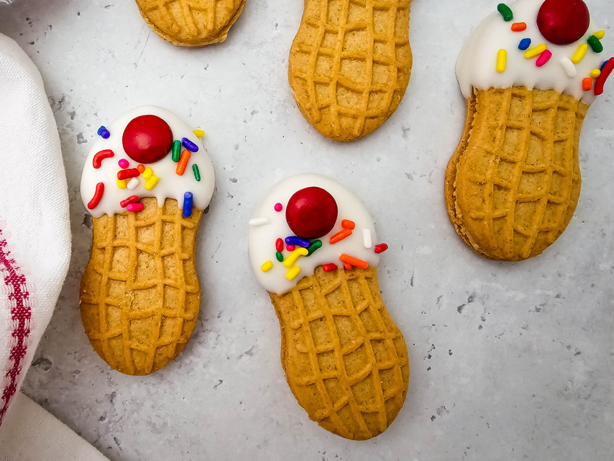 A close-up shot focusing on three decorated peanut butter sandwich cookies styled like mini ice cream sundaes with white candy coating, colorful sprinkles, and a red candy on top, sitting on a light gray background.