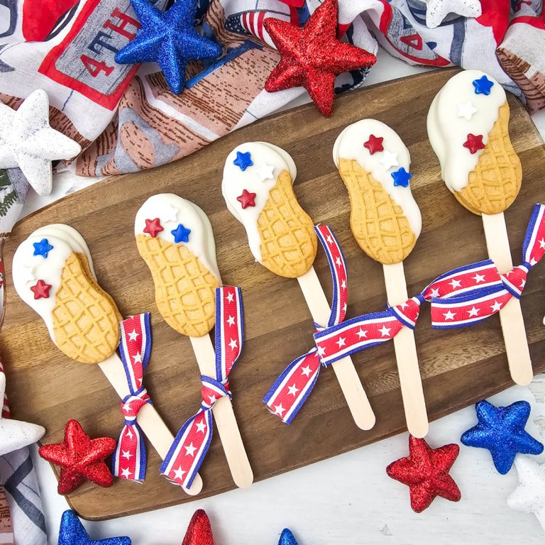 A festive overhead feature shot showing a collection of patriotic nutter butter cookie pops on a rustic wooden board. The treats are decorated with smooth white chocolate and red, white, and blue star sprinkles, surrounded by sparkling holiday star ornaments.