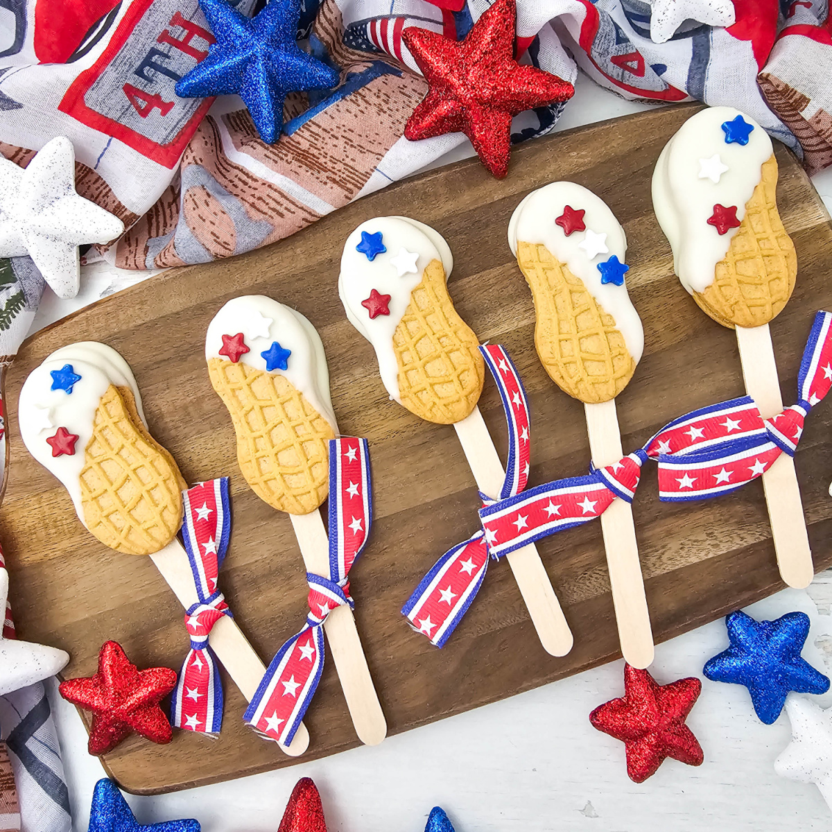 A festive overhead feature shot showing a collection of patriotic nutter butter cookie pops on a rustic wooden board. The treats are decorated with smooth white chocolate and red, white, and blue star sprinkles, surrounded by sparkling holiday star ornaments.