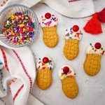 A wide, top-down shot of five decorated peanut butter cookies made to look like mini ice cream cones, displayed on a light surface next to a clear bowl filled with rainbow sprinkles and a white dish towel featuring a red border and tassel.