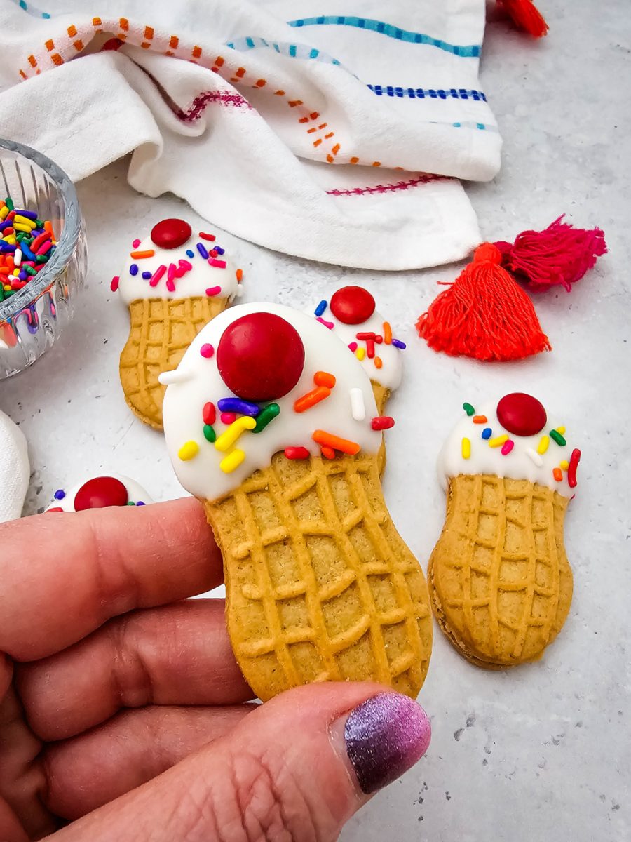 A close-up view of a hand holding up a decorated peanut butter sandwich cookie made to look like a mini ice cream cone, with other decorated cookies and a bowl of sprinkles blurred in the background.