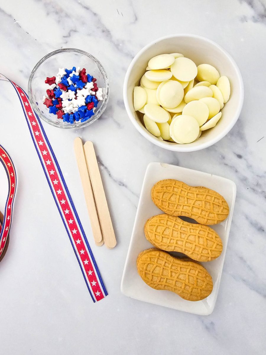 Ingredients laid out on a marble countertop to make patriotic nutter butter cookie pops, including a bowl of white melting chocolate wafers, Nutter Butter cookies, red, white, and blue star sprinkles, and popsicle sticks.