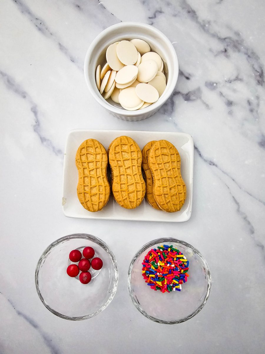 A top-down overhead view of the ingredients needed for the recipe, including a white bowl of white melting wafers, a small plate with three peanut butter sandwich cookies, a small glass bowl with red chocolate candies, and a small glass bowl with rainbow sprinkles, all arranged on a white marble countertop.